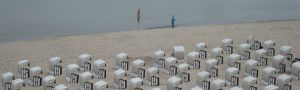 Das Bild zeigt einen Strand mit zahlreichen weißen Strandkörben, die in geordneter Weise auf dem Sand aufgestellt sind. Im Hintergrund sind zwei Personen am Wasser zu sehen, die den ruhigen Strand entlanggehen. Das Meer wirkt still, und die Szene vermittelt eine ruhige, fast meditative Atmosphäre. Der Fokus liegt auf der Struktur der Strandkörbe und dem weiten, leeren Raum am Strand.